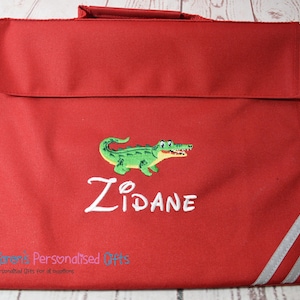 May include: Red book bag with a white embroidered alligator and the name "ZIDANE" on the front. The bag has two white stripes on the right side.