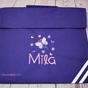 Puede incluir: Mochila de color morado con rayas blancas a los lados. La mochila tiene un bordado de mariposa y flores con el nombre "Mila" en rosa.