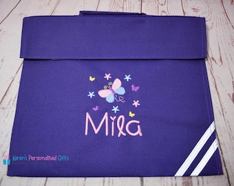 May include: Purple book bag with white stripes on the sides. The bag has a butterfly and flower embroidery with the name "Mila" in pink.