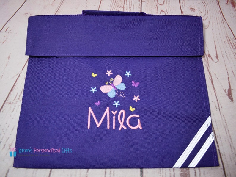 Puede incluir: Mochila de color morado con rayas blancas a los lados. La mochila tiene un bordado de mariposa y flores con el nombre "Mila" en rosa.