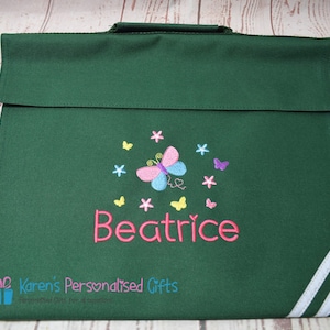 Puede incluir: Bolso de libros verde con rayas blancas en el lateral. El bolso tiene una mariposa con alas rosas, azules y amarillas y el nombre "Beatrice" bordado en la parte delantera.