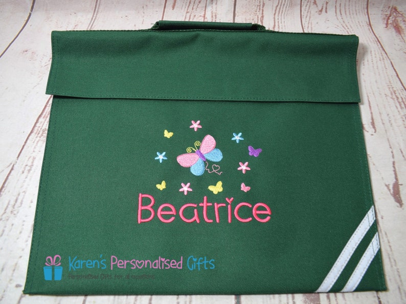 Puede incluir: Bolso de libros verde con rayas blancas en el lateral. El bolso tiene una mariposa con alas rosas, azules y amarillas y el nombre "Beatrice" bordado en la parte delantera.