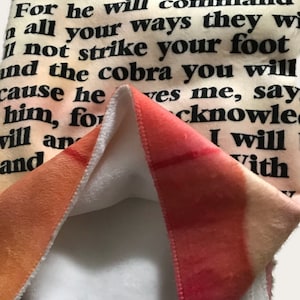 Puede incluir: Una manta de forro polar suave y blanca con un diseño degradado de colores en la parte posterior. La parte delantera de la manta presenta una impresión de texto en blanco y negro de un versículo bíblico: "El Señor es mi refugio, ningún mal te alcanzará. Porque él mandará a todos tus caminos, no tropezarás con tu pie y la cobra no te picará."