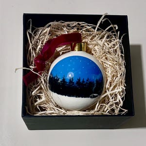 Peut inclure: Une boule de Noël blanche avec une scène nocturne peinte d'une forêt sous un ciel étoilé. L'ornement a un capuchon doré et un ruban de velours rouge. Il est présenté dans un coffret cadeau noir avec de la paille.