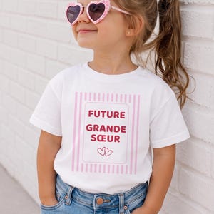 Peut inclure: Un t-shirt blanc à manches courtes avec un cadre rectangulaire à rayures roses, portant l'inscription FUTURE GRANDE SŒUR en lettres rouges et deux petits cœurs rouges, associé à un jean bleu et des lunettes de soleil roses en forme de cœur.