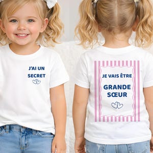 Peut inclure: Un t-shirt blanc à manches courtes avec le texte bleu J'AI UN SECRET et deux cœurs sur le devant. Le dos présente un rectangle à rayures roses avec le texte JE VAIS ÊTRE GRANDE SŒUR et deux cœurs en dessous.