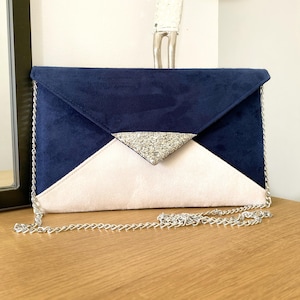 Op de afbeelding: Een marineblauwe en crèmekleurige clutch met een zilveren glitter driehoek accent. De clutch heeft een zilveren kettingriem en een geometrisch ontwerp. De tas is gemaakt van een zacht, suède-achtig materiaal.