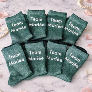 Peut inclure: Huit paires de chaussettes vertes scintillantes avec l'inscription "Team Mariée" en blanc. Les chaussettes sont pliées et disposées sur une surface en marbre, avec des perles et des pétales de rose.