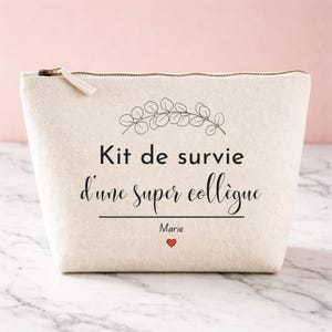 Peut inclure: Trousse de maquillage en toile beige avec fermeture éclair dorée. Le texte "Kit de survie d'une super collègue" est écrit en noir, avec le nom "Marie" et un cœur rouge en dessous. Une branche de feuilles est au-dessus.