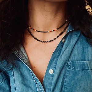 May include: A person wearing a denim shirt with two layered necklaces. The top necklace is a thin black beaded necklace with colorful beads. The bottom necklace is a thicker black beaded necklace.