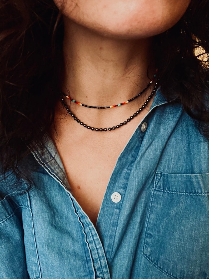 May include: A person wearing a denim shirt with two layered necklaces. The top necklace is a thin black beaded necklace with colorful beads. The bottom necklace is a thicker black beaded necklace.