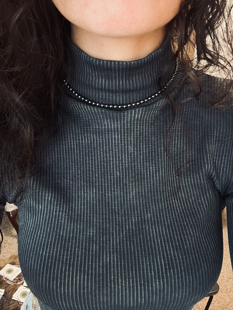 May include: A close-up of a person wearing a dark blue turtleneck sweater with a silver beaded necklace.