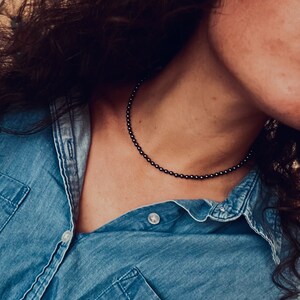 May include: A person wearing a denim shirt with a black beaded necklace.