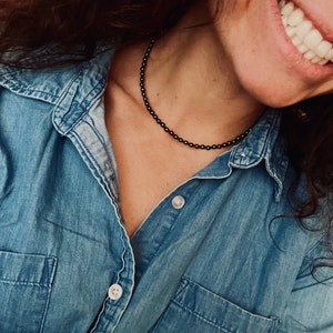 May include: A woman wearing a denim shirt with a black beaded necklace.