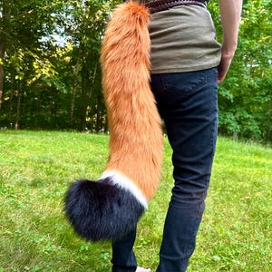 May include: A faux fur fox tail, primarily orange with a white and black tip, attached to a belt. The tail is long and fluffy, designed to mimic a fox's tail. The person is wearing dark blue jeans and a green shirt.