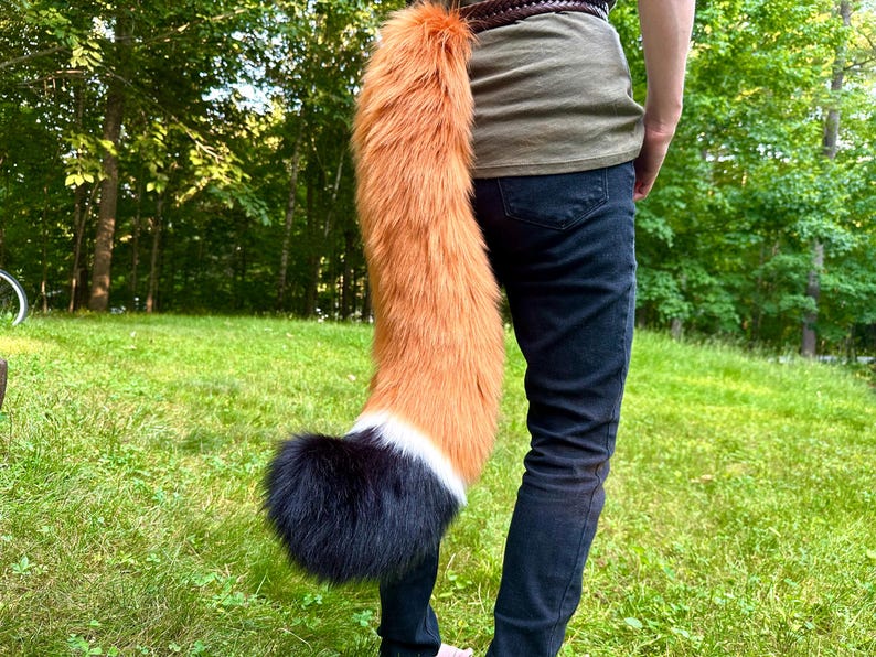 May include: A faux fur fox tail, primarily orange with a white and black tip, attached to a belt. The tail is long and fluffy, designed to mimic a fox's tail. The person is wearing dark blue jeans and a green shirt.