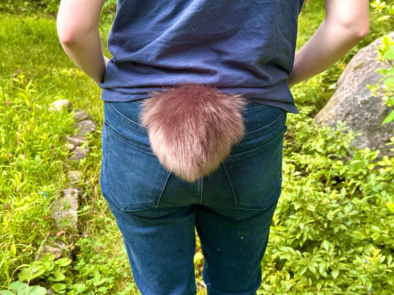 Peut inclure: Une queue en fausse fourrure duveteuse, brun rouge&acirc;tre, attach&eacute;e &agrave; l'arri&egrave;re d'un jean bleu. La queue est un accessoire de costume. L'arri&egrave;re-plan est un cadre ext&eacute;rieur naturel avec du feuillage vert.