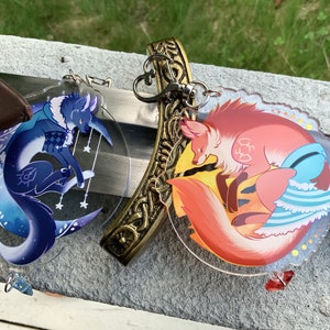 May include: Two acrylic keychains featuring colorful illustrations of foxes. One keychain depicts a blue fox with a crescent moon and stars, while the other keychain shows a red fox with a sun and clouds.