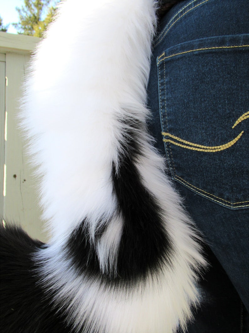 May include: A black and white faux fur tail with a white tip. The tail is attached to a person's waist and is hanging down.
