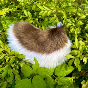 以下が含まれることがあります： ふわふわの茶色と白のフェイクファーのアイテム。ペット用ベッドやアクセサリーかもしれません。鮮やかな緑の葉の中にあります。アイテムは丸みを帯びた形で、茶色の中心と白い縁があります。水色のリボンが付いています。