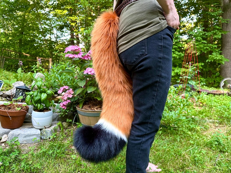 May include: A faux fox tail in shades of orange, white, and black. The tail is attached to a brown belt and hangs down. The tail is fluffy and appears to be made of faux fur. The person is wearing dark blue jeans.