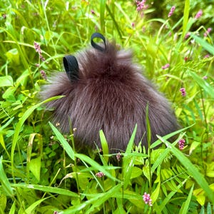 Peut inclure: Un accessoire brun fonc&eacute; et duveteux avec deux boucles noires, nich&eacute; dans une herbe verte vibrante et de petites fleurs violettes. L'article a un aspect doux et textur&eacute;, contrastant avec l'environnement naturel. L'accessoire est probablement un article de mode.
