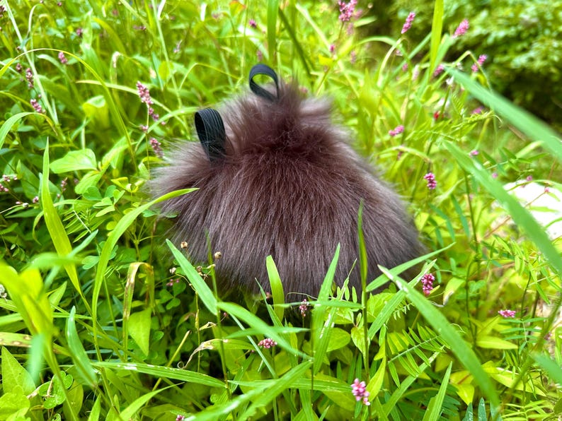 Peut inclure: Un accessoire brun fonc&eacute; et duveteux avec deux boucles noires, nich&eacute; dans une herbe verte vibrante et de petites fleurs violettes. L'article a un aspect doux et textur&eacute;, contrastant avec l'environnement naturel. L'accessoire est probablement un article de mode.