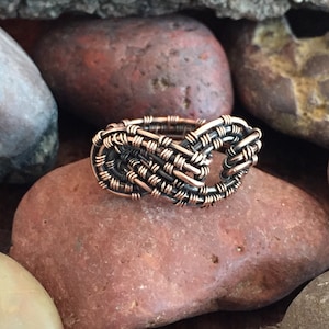 May include: A copper and silver wire ring with a woven design. The ring is shaped like an infinity symbol.