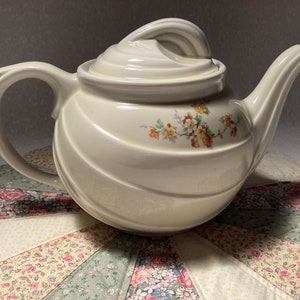May include: A white ceramic teapot with a floral design. The teapot has a round body with a swirling pattern and a lid with a knob on top. The teapot is sitting on a colorful patchwork fabric.