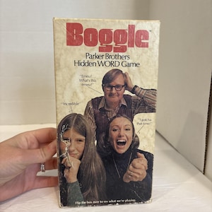 Pode incluir: Caixa de jogo Boggle vintage com a palavra "Boggle" em vermelho. A caixa apresenta uma imagem retrô de três pessoas com balões de fala. O texto "Parker Brothers Hidden WORD Game" também é visível.