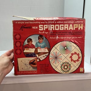 May include: Vintage Spirograph drawing toy box. The red box features the text "Kenner's NEW SPIROGRAPH" and illustrations of children using the toy. The box also shows examples of the patterns that can be created.