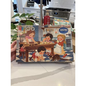 Puede incluir: Caja de rompecabezas vintage MB 100 Little Ben con una colorida ilustración de niños horneando galletas. La caja tiene un patrón de rayas azules y blancas y muestra el título y la marca del rompecabezas.