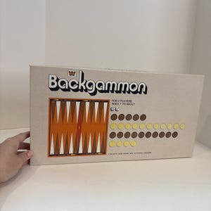 Puede incluir: Un juego de backgammon en caja. La palabra "Backgammon" está escrita en negro en la caja. El tablero es naranja y marrón, con fichas marrones y amarillas. El texto de la caja dice "FOR 2 PLAYERS AGES 7 TO ADULT".