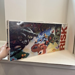 May include: Vintage Risk board game box. The box features a world map with territories and a cannon illustration. The word "RISK" is prominently displayed in red. The game is a Parker Brothers World Conquest Game.