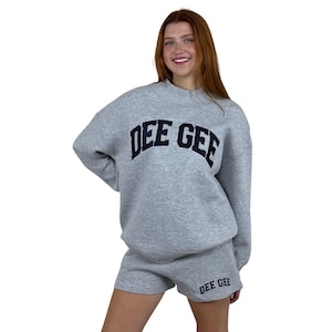 May include: A light gray long-sleeve sweatshirt and matching shorts set. The sweatshirt features a mock neck and the text DEE GEE in dark navy blue block letters across the chest. The shorts also feature the text DEE GEE in navy blue on the leg.