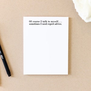 May include: A white notepad with the text "Of course I talk to myself... sometimes I need expert advice."  Two black pens are on a brown surface next to the notepad.