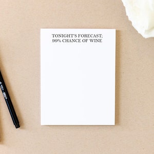 May include: A white notepad with the text "TONIGHT'S FORECAST; 99% CHANCE OF WINE" printed on the top. Two black pens are lying on a brown surface next to the notepad.