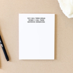 May include: A white notepad with the text "You call them swear words. I call them sentence enhancers." printed on the top. Two black pens are lying on a brown surface next to the notepad.
