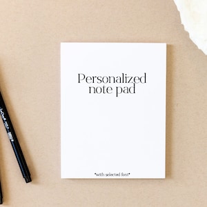 May include: A white notepad with the text "Personalized note pad" and "*with selected font*" printed on it. Two black pens are lying on a brown surface next to the notepad.