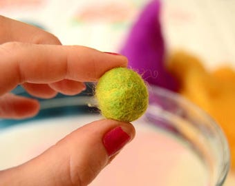 May include: A small, round, lime green felt ball is held between two fingers. The felt ball is a vibrant green with yellow highlights. In the background, a purple felt cone and a yellow felt shape are visible.