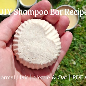 以下が含まれることがあります： 手で持たれた、ハート型のデザインの明るい色の丸いシャンプーバー。バーは、小さな容器に入った他の材料に囲まれています。画像には「DIY Shampoo Bar Recipe」と「For Normal Hair | Nettle & Oat | PDF Guide」と書かれています。