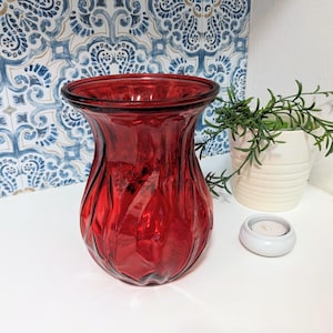 May include: A ruby red glass vase with a textured, fluted design. The vase is empty and sits on a white surface, with a small white candle holder and a potted plant in the background. The vase has a wide opening.