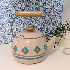 May include: A cream-colored enamel teapot with a wooden handle and lid knob. The teapot features a design of blue and orange diamonds and stripes. The spout and handle are silver. The teapot is approximately 8 inches in height.