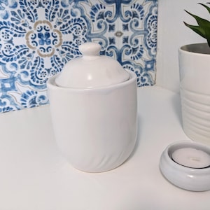 May include: White ceramic jar with a lid, a white ceramic candle holder, and a small white candle. The jar and candle holder are on a white surface with a blue and white patterned tile background.