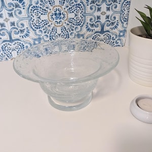 May include: A clear glass bowl with a textured surface and a wide rim. The bowl is sitting on a white surface.