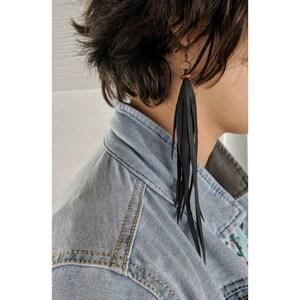 May include: A person wearing a denim jacket with a long black leather earring. The earring is made of thin strips of leather that hang down from a small metal ring.