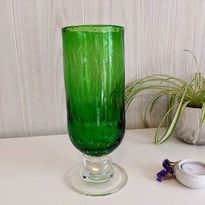 May include: A tall, green glass vase with a textured surface and a clear glass base. The vase is sitting on a white surface.