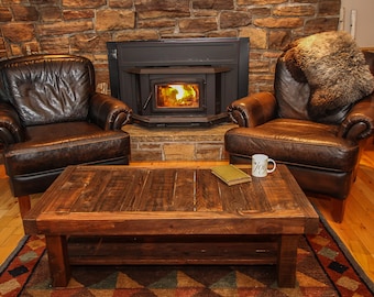 Può includere: Due poltrone in pelle marrone con un tavolino da caffè in legno davanti a un camino in pietra con una stufa a legna. Il tavolino ha una venatura del legno rustica e uno scaffale inferiore. Un libro e una tazza sono sul tavolo.