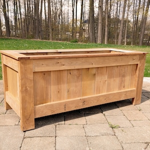 Könnte beinhalten: Eine rechteckige Pflanzkasten aus hellfarbenem Holz. Der Pflanzkasten hat ein schlichtes, rustikales Design mit vertikalen Brettern an den Seiten und einer rechteckigen Öffnung oben. Der Pflanzkasten steht auf einer Steinterrasse.