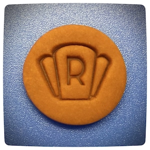 May include: A round brown cookie with a raised design of a letter "R" inside a square shape with three curved lines on each side.